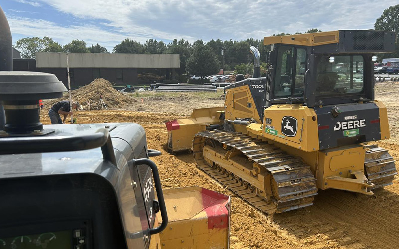 John Deere 700L bulldozer operating on a construction site with a worker in the background.