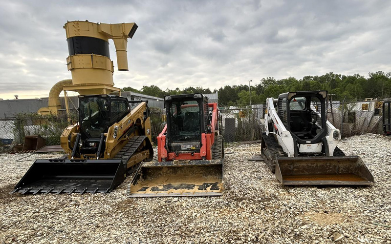 Three skid steer loaders in different colors parked on gravel at a construction site.